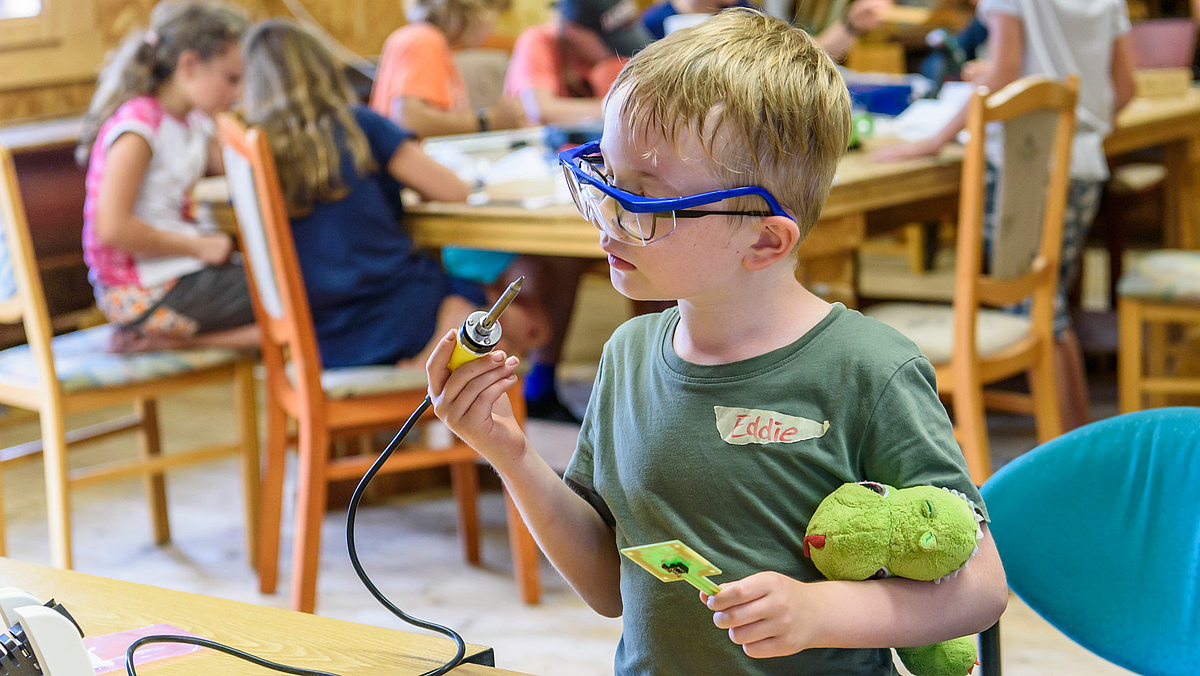 Das Foto zeigt Eddie, einen kleinen Jungen mit einem Dino-Kuscheltier unter dem linken Arm. Er betrachtet genau den Lötkolben, den er in der rechten Hand hält.