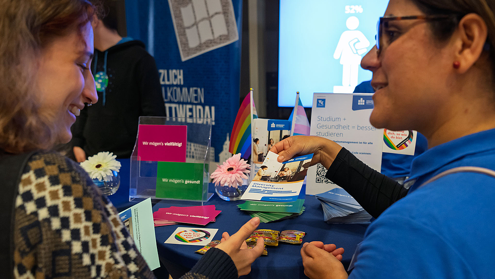 Weibliche Person überreicht an andere weibliche Person Flyer mit der Überschrift "Diversity Management".