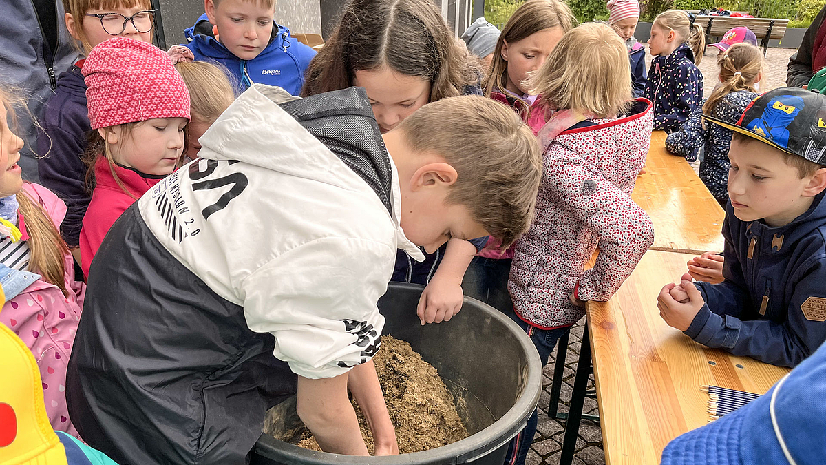 Zahlreiche Kinder stehen um einen großen Bottich mit Erde. Ein Kind hat seine Hände im Bottich.