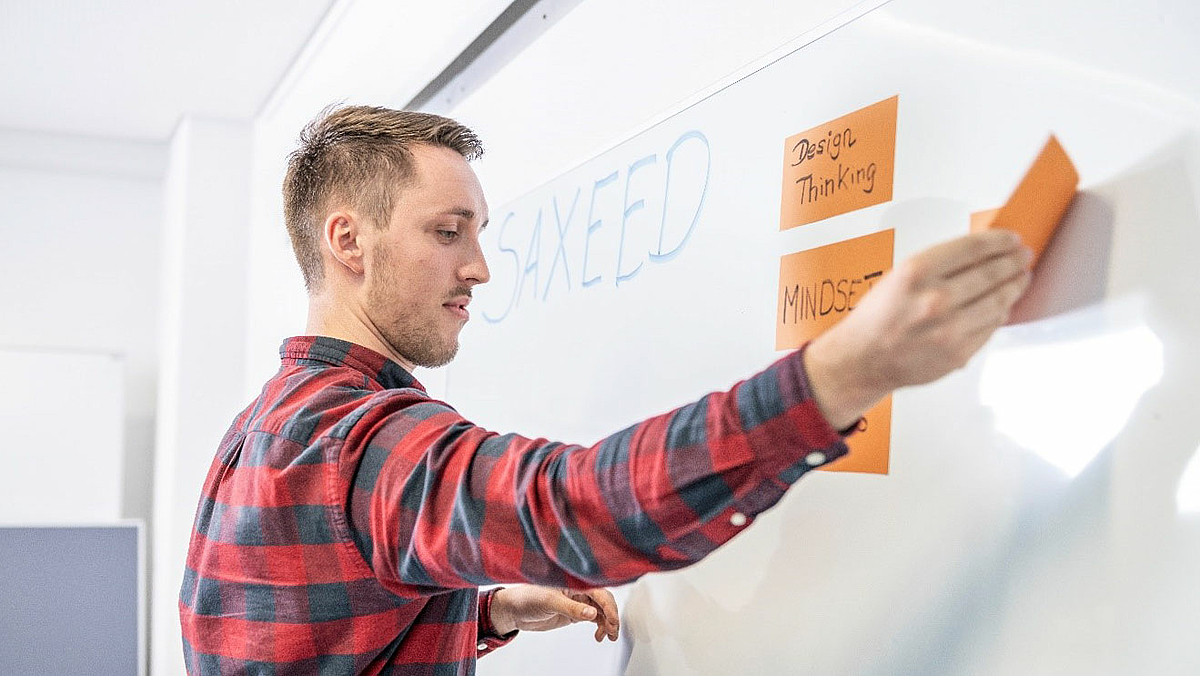 Das Foto zeigt eine junge männliche Person, die beschriftete Kärtchen an ein Whiteboard heftet, auf dem groß der Schriftzug SAXEED geschrieben ist. Auf den Moderationskarten ist einmal handgeschrieben "Design Thinking" und einmal "Mind Set" zu lesen.