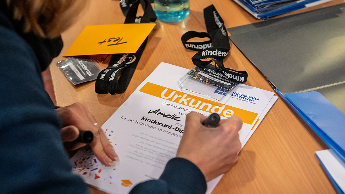 Person schreibt den Namen "Amelie" auf eine Urkunde. Auf dem Tisch liegen mehrere Mappen, dunkelblaue Schlüsselbänder mit weißer Aufschrift „Kinderuni“.