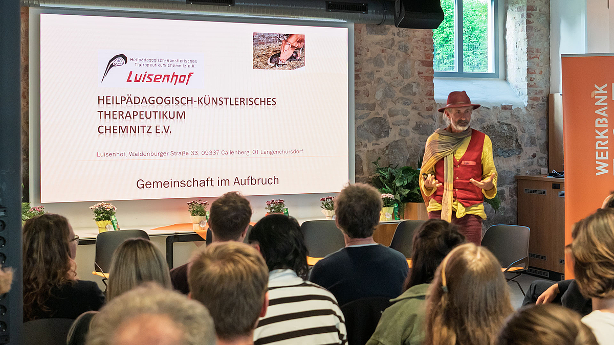 Vom Bauingenieur zum Mitgründer der Luisenhof-Gemeinschaft: Robby Hammer erzählt die Geschichte des Hofs bis zur ausgezeichneten Institution für Inklusionsarbeit mit Jugendlichen. Eine männliche Person mit Hut spricht zum Publikum. Auf dem Screen links neben ihr ist unter anderem zu lesen „Luisenhof. Gemeinschaft im Aufbruch“.