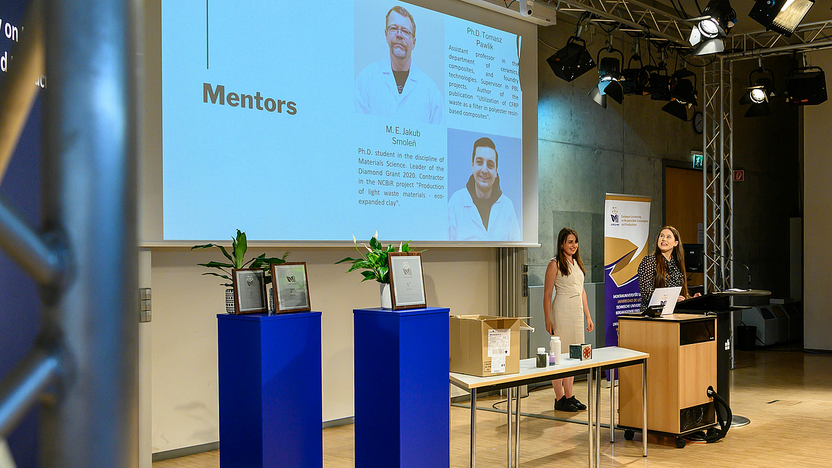 Zwei junge Frauen am Stehpult im Hintergrund präsentieren. Auf der Leinwand im Bild links neben ihnen ist eine Folie mit Porträts zweier männlicher Personen und dem Titel „Mentors“ zu sehen.