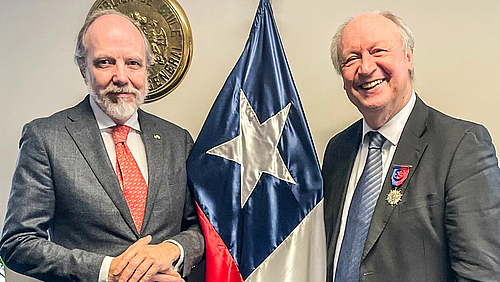 Der chilensiche Generalkonsul Matías Undurraga Abbott und der soeben mit dem "Orden al Mérito Bernardo O'Higgins" ausgezeichnete Mittweidaer Professor Hans-Peter Niedermeier (v.l.) Das Foto zeigt zwei ältere männliche Personen. Ziwichen ihnen ist senkrecht die chilensiche Nationalflagge zu sehen. Die erchte Person trägt an der linke Brust den Orden am Band - ein silbervergoldeter fünfstrahliger Stern mit einer weiß emaillierten Fassung, der auf einem ebenfalls fünfstrahligen Stern versetzt aufliegt.