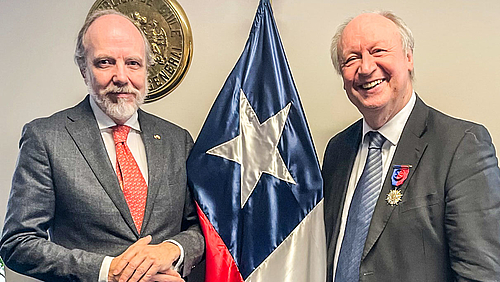 Der chilensiche Generalkonsul Matías Undurraga Abbott und der soeben mit dem "Orden al Mérito Bernardo O'Higgins" ausgezeichnete Mittweidaer Professor Hans-Peter Niedermeier (v.l.) Das Foto zeigt zwei ältere männliche Personen. Ziwichen ihnen ist senkrecht die chilensiche Nationalflagge zu sehen. Die erchte Person trägt an der linke Brust den Orden am Band - ein silbervergoldeter fünfstrahliger Stern mit einer weiß emaillierten Fassung, der auf einem ebenfalls fünfstrahligen Stern versetzt aufliegt.