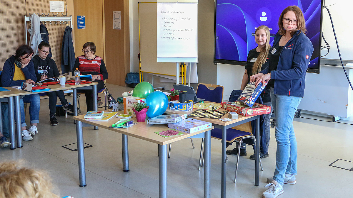 Zwei jüngere weibliche Personen stehen in einem Seminarraum hinter einem Tisch, auf dem verschiedenste Gegenstände liegen. Im Hintergrund sitzen weitere weibliche Pesonen an dienem Tissch