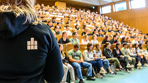 Voller Hörsaal zum Studienstart, aber noch ist Platz in ausgewählten Studiengängen. Die Bankreihen eines Hörsaals sind voll besetzt. Am linken Bildrand steht eine Person, die sich den Pesonen im Hörsaal zuwendet. Sie trägt einen blauen Kapuzenpullover mit dem Hochschullogo auf dem Rücken.