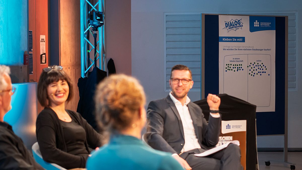 Im Vordergrund unscharf das Podium. Im Hintergrund scharf eine Stellwand mit dem Text: "Umfrage zur heutigen Veranstaltung: Wo würden Sie Ihren nächsten Staubsauger kaufen?" und aufgeklebten Punkten in zwei Feldern. Gegenüber dem Feld "lokal im Geschäft" hat das Feld "online" mehr Punkte.