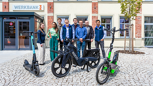 Das Team des Instituts für Energiemanagement der Hochschule Mittweida (ifem) und ihrer Kooperationspartner:innen mocci und blXmove stehen vor der Zukunft vernetzter Mobilität in der Hochschulstadt. (Foto: Erik Hilse) Das Foto zeigt eine Gruppe von 8 Personen auf einem gepflasterten Platz vor einem Gebäude stehend, das den Namen Werbank32 trägt. Vor der Gruppe stehen drei zweirädrige Fahrzeuge, zwischen zwei E-Scootern ein futuristisches Pedelec mit der Aufschrift mocci.