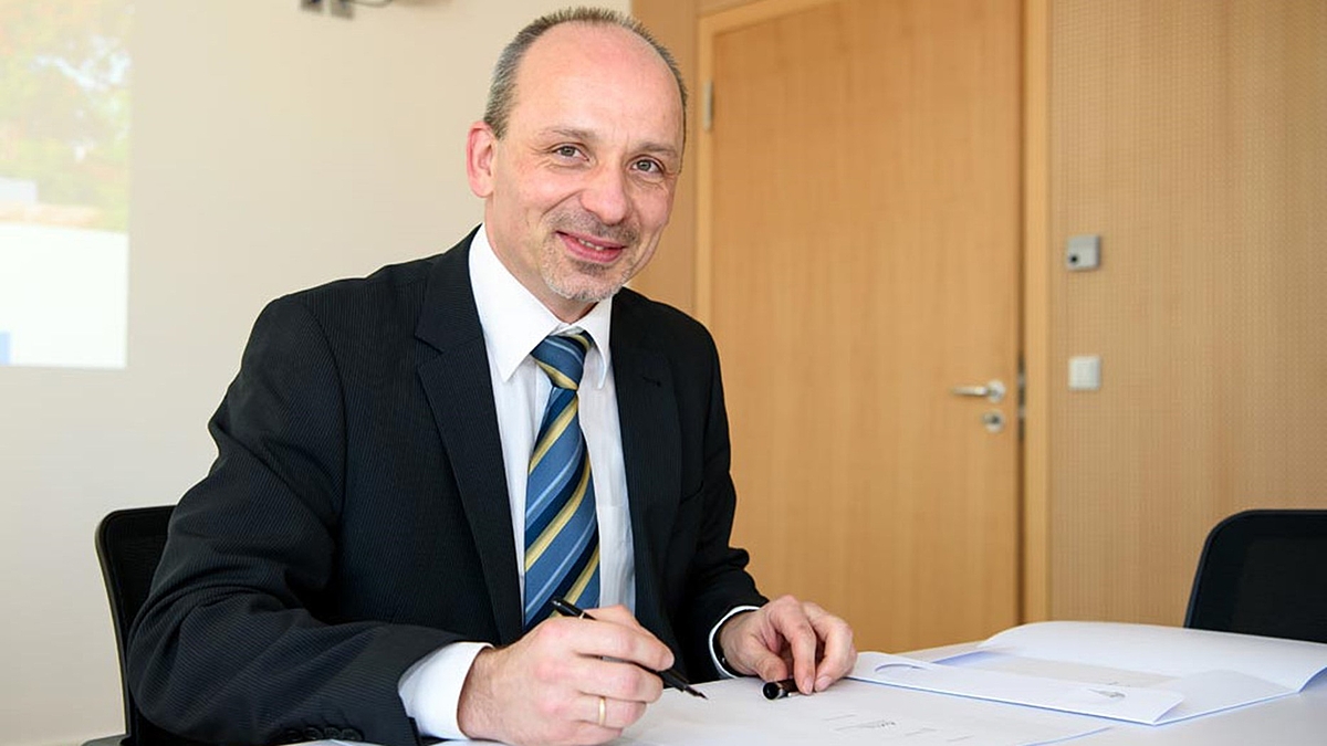 Jörg Matthes sitzt in dunklem Anzug mit blau-gold-schwarz gestreifter Krawatte an einem Besprechungstisch. In der rechten Hand hält er einen Füllfederhalter, auf dem Tisch liegen Dokumente.