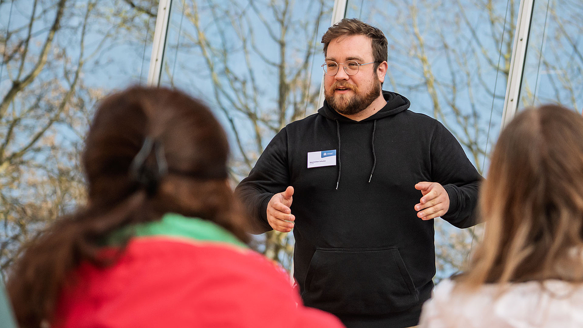 Eine junge mänliche Person steht gestikulierend vor einer Glasfront. Im Bildvordergrund sind Personen von hinten zu sehen.