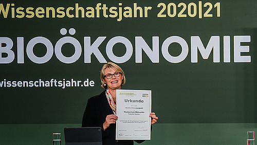Das Foto zeigt eine lächelnde weilbliche Person, die eien Urkunde in Richtung Kamera hält. Auf der Urkunde ist u.a. "Hochschule Mittweida" zu lesen. Im Hiuntergrund sich groß die Wörter "Wissenschaftsjahr 2020/21 Bioökonomie" zu lesen.  