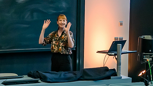 Zeiten wenden! Die Journalistin und Sachbuchautorin Dr. Susanne Götze blickt in der Ringvorlesung an der Hochschule Mittweida auf die Bremser beim Wandel in der Klimakrise. Blick auf die Dozentin vorne am Pult, hinter ihr die Tafel. Sie hält ihre Hände wie abwehrend nach oben, blickt aber lachend ins (nicht sichtbare) Publikum.