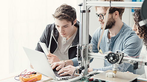 Zwei junge Männer sitzen vor einem Laptop. Neben ihnen steht ein 3D-Drucker, der ein Plastikteil herstellt.