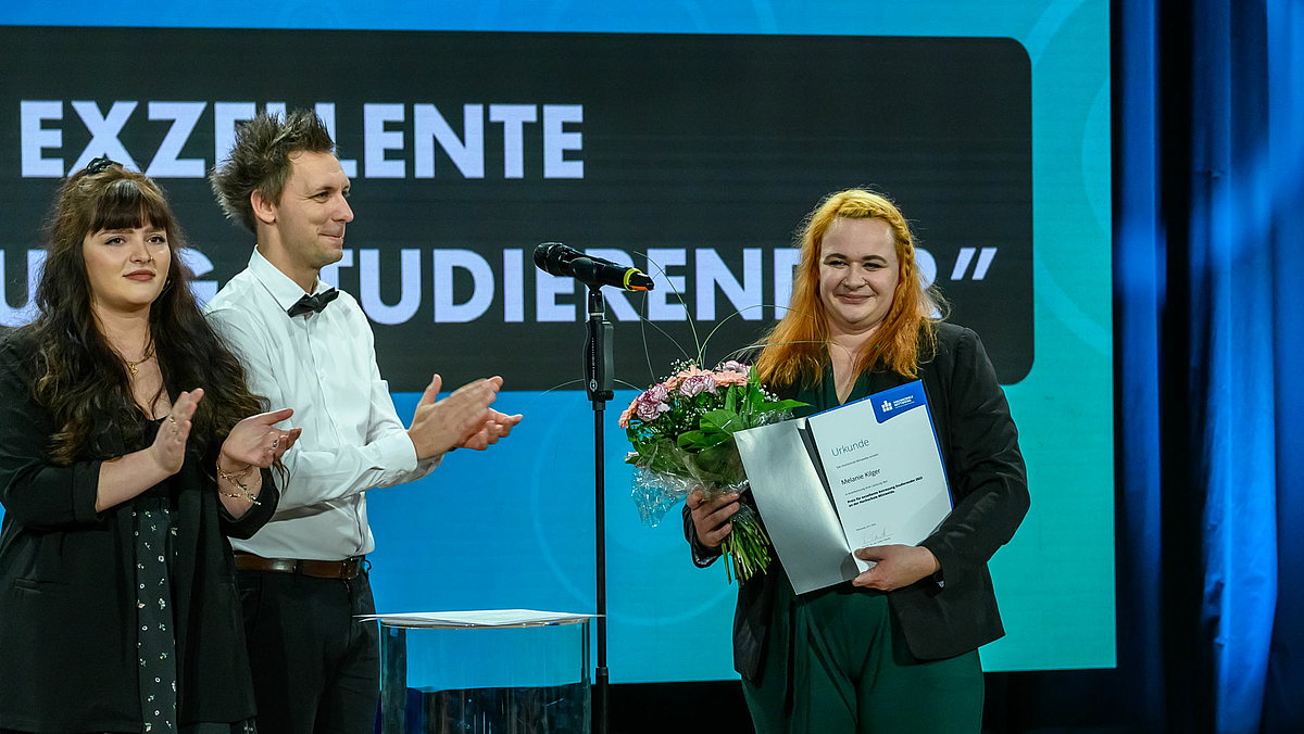 Selbe Kulisse wie zuvor. Ein männliche und zwei weibliche Personen stehen vor dem Screen. Die weibliche Person rechts hält Blumen und Urkunde in den Händen.