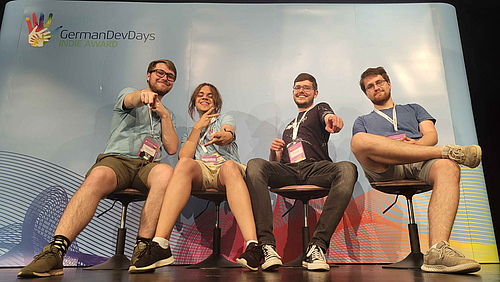 Das Team von Traveller's Box hat sich bereits auf Branchen-Events wie den German Dev Days und der Gamescom einen Namen gemacht: Laurenz Bischoff, Robin Schöppach, Raphael Müller) und Johannes Bätz (v.l.). Vier junge Menschen sitzen fröhlich gestikulierend vor eine Messewand mit dem Schriftzug „German DevDays. INDIE AWARD“.