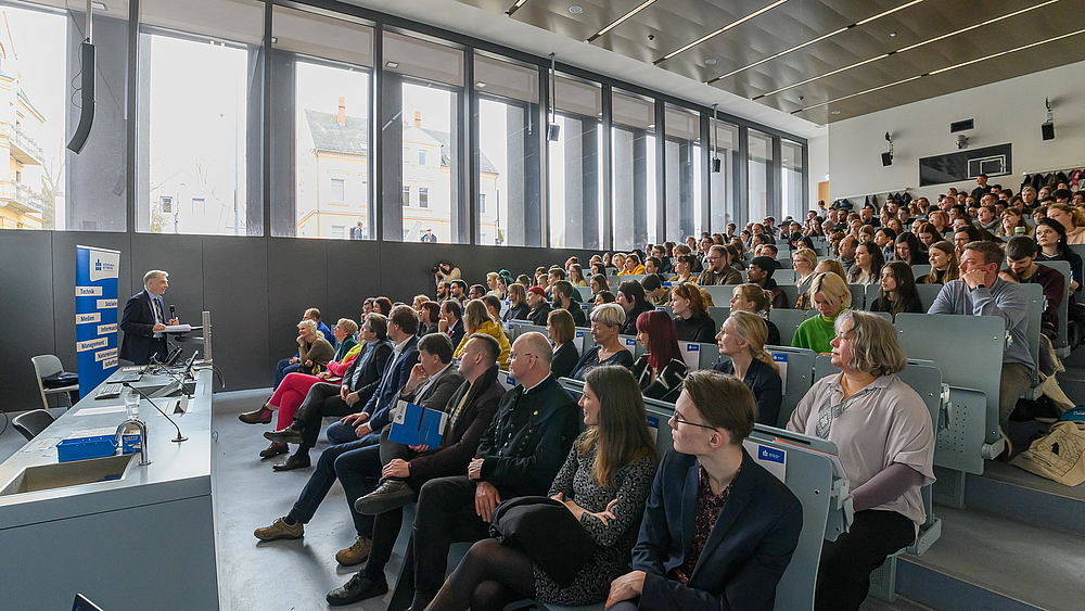 Der Rektor feierlich: „Ich immatrikuliere Sie in die Gemeinschaft der Hochschule Mittweida.“ Seitlicher Blick in den vollbesetzten Hörsaal. Links steht eine männliche Person an einem Pult, richtet sich an das Plenum.