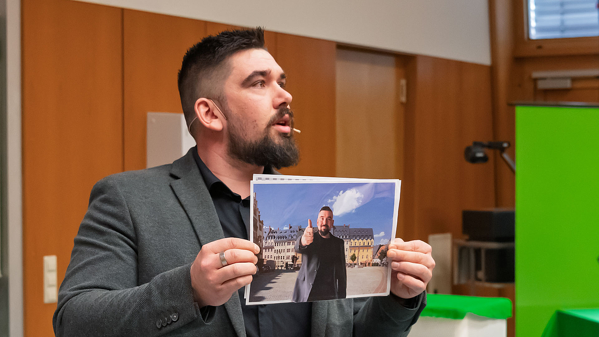 Der Dozent zeigt den Kindern ein Foto, das ihn vor einem künstlichen Hintergrund zeigt, , Im Hintergrund sind eine grüne Wand und ein Tisch mit grünem Stoff zu sehen.