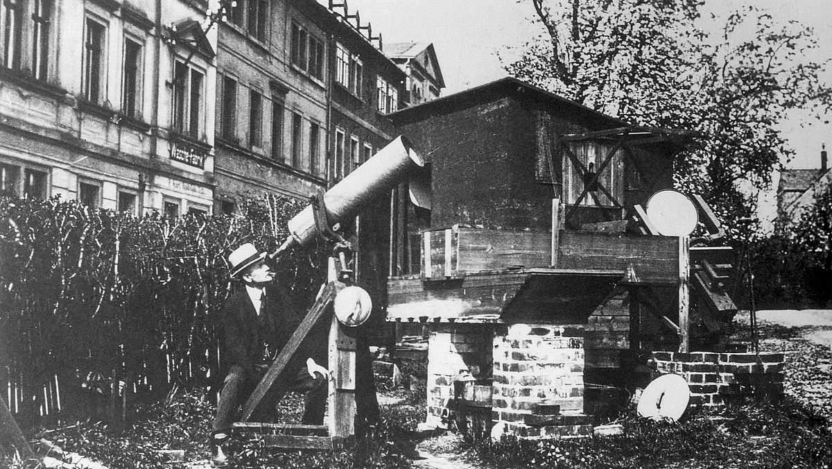 Die Schwarzweiß-Aufnahme zeigt einen Mann mit Hut in einem Garten, der durch ein, auf einem hölzernen Stativ montiertes, großes Teleskop schaut. Die Blickrichtung des Teleskops ist als Gegenschuss zum Aufmacherfoto mit den sich spiegelnden Objektivlinsen zu interpretieren.