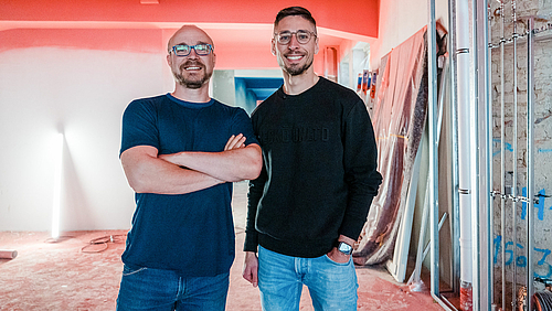 Gemeinsam für die Games-Branche in Mitteldeutschland: Auf der Baustelle von R42 treffen sich der Mittweidaer Professor Alexander Marbach und der Mitbegründer von R42 Tom Potutschek (v.l.). (Foto: GECKO.2) Zwei männliche Personen stehen strahlend lächeln nah beeinander. Die linke Person hat die Arme verschränkt. Im Bildhintergrund ist dutliche zu erkennen, dass sie sich in einem Gebäude befinden, in dem gerade der Innenausbau stattfindet.