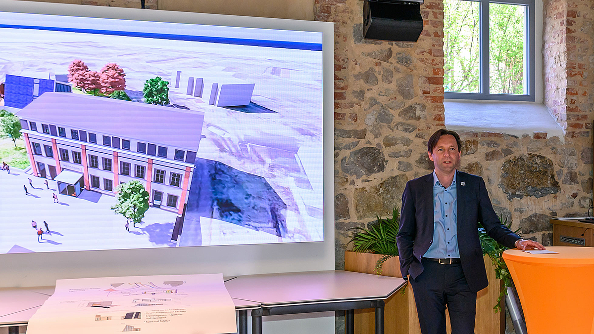 „Mittweida ist ein Paradebeispiel für innovationsgestützte Regionalentwicklung“, lobt Ronny Zienert vom SMR die Zusammenarbeit von Wissenscha Das Foto zeigt eine männliche Person an einem Stehtisch. Auf dem Bild links daneben ist auf einem großen Display die Animation des Werkbank32-TELEWERK-Geländes zu sehen.