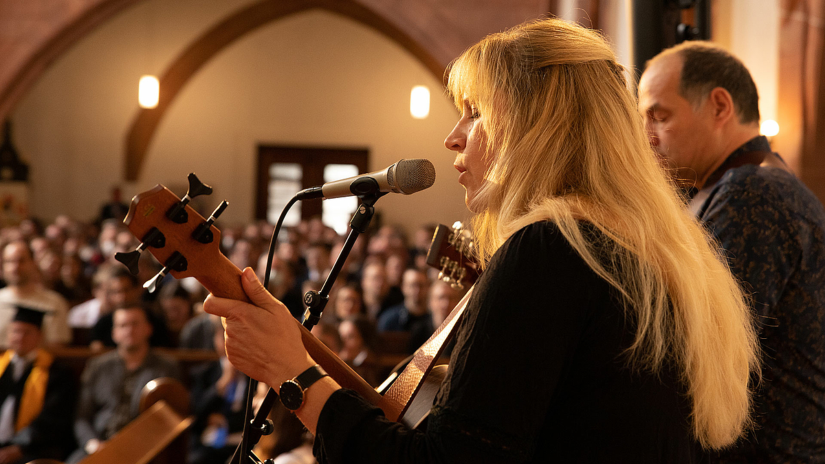 Eine Frau mit Gitarre singt ruhig in ein Mikrofon. Rechts neben ihr steht ein Mann. Im Hintergrund sitzen die Studierenden mit ihren Eltern auf den Kirchenbänken und lauschen.