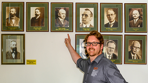 That’s him: Urgroßvater Julius Anselm in der Galerie der früheren Direktoren und Rektoren der Hochschule Mittweida mit Urenkel Prof. Martin Anselm vom Rochester Institute of Technology. Eine männliche Person blickt lächelnd in die Kamera. Mit dem rechten Zeigefinger zeigt sie auf das Schwarzweiß-Porträt eines Mannes, das neben weiteren Porträts in gleichen schlichten Holzrahmen in zwei Riehen an der Wand im Hintergrund aufgehängt ist.