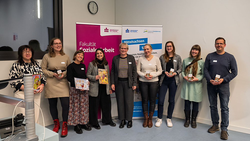 Gruppe aus acht weiblichen und einer männlichen Person am unteren Ende des Hörsaals vor Rollups mit "Fakultät Soziale Arbeit" und "Mittelsachsen Sozial"