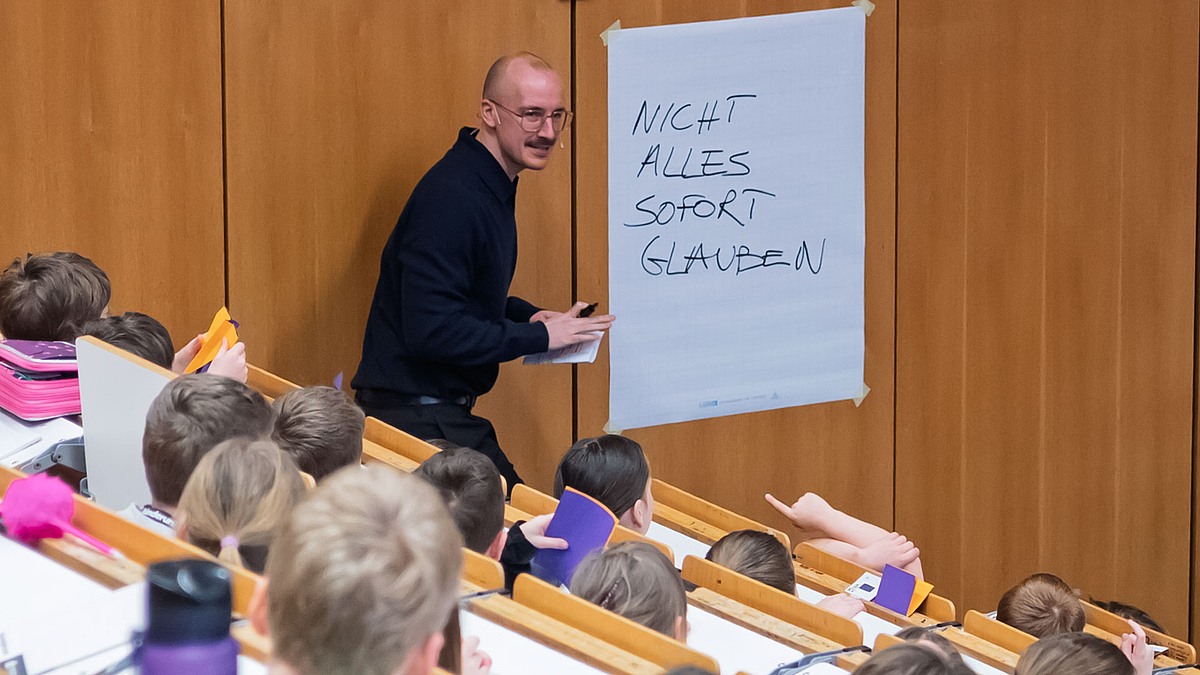 Der Dozent steht vor einem Blatt Papier, das an der Wand befestigt wurde. Auf das Papier hat er gerade geschieben: „Nicht alles sofort glauben“.