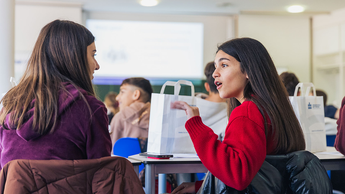 Zewei Mächsen in einem Seminaraum unterhalten sich. Vor ihnen auf dem Tisch stehen Papiertüten mit HSMW-Logo