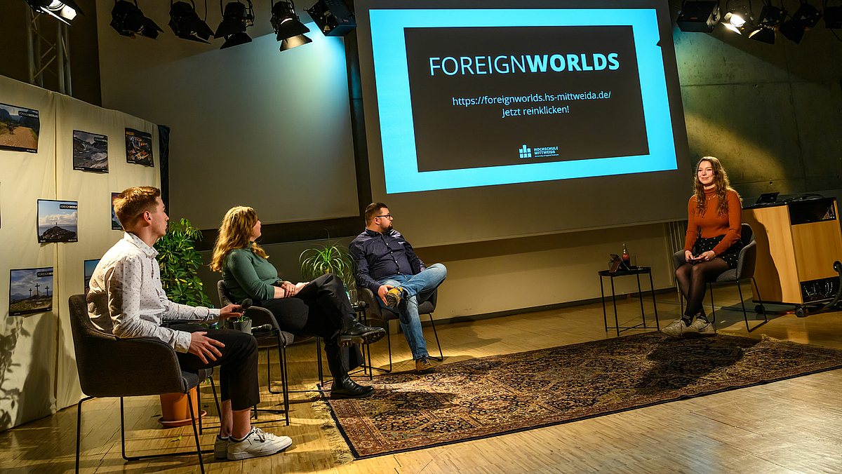 Das Foto zeigt die studentische Talkrunde aus dem Aufmacherfoto. Auf der Leinwand im Hintergrund ist zu lesen: „FOREIGNWORLDS https://foreignworlds.hs-mittweida.de Jetzt reinklicken!“