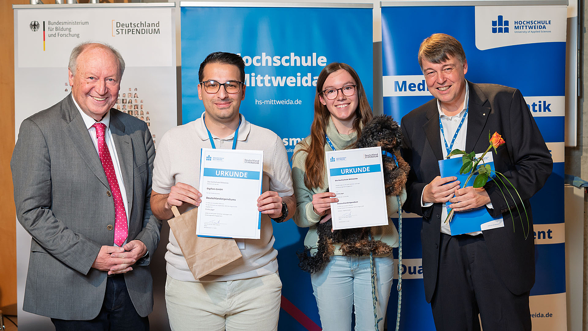 Eine männliche und eine weibliche Person mit Urkunden zwische den beiden Urkundenübergebenden vor dem bekannten Hintergrund.