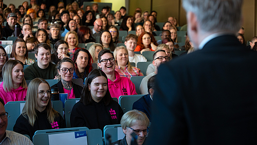 Feierlicher Akt, gelöste Stimmung. Rektor Volker Tolkmitt nahm die neuen Studierenden bei der Immatrikulationsfeier in die Hochschulgemeinschaft auf - und sorgte für Unterhaltung. Im Vordergrund steht ein Mann mit grauen Haaren im Anzug, der von hinten zu sehen ist. Vor ihm sitzen auf den Reihen eines Hörsaals viele Studierende, die vergnügt lachen.