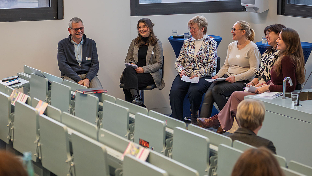 Eine männliche und fünf weibliche Personen auf Hockern am unteren Ende des Hörsaals.