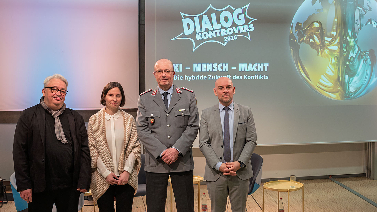 Vier Personen, darunter eine weibliche, stehen nebeneinander für ein Gruupenfoto. Auf der Leinwand im Hintergrund ist das Dialog-Kontrovers-Logo und der Titel der aktuellen Veranstaltung zu lesen: „KI, Mensch & Macht – Die hybride Zukunft des Konflikts“