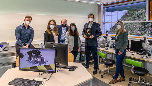 Das Foto zeigt drei junge Frauen und drei junge Männer in einem Labor mit Mikroskopen im Hintergrund. Alle tragen FFP2-Masken. Auf einem Tisch vor der Personengruppe ist auf einem PC-Monitor eine Grafik mit dem Schriftzug 3D-FORCE zu sehen.