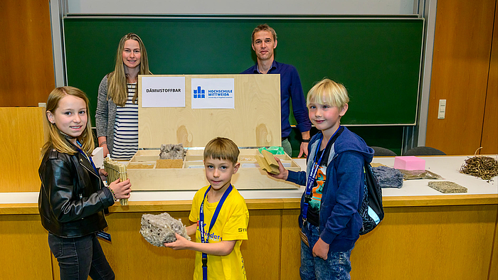 Das Foto zeigt im Vordergrund drei Kinder (ein Mädchen, zwei Jungs) vor einem Hörsaalpult auf dem ein Kasten mit der Aufschrift Dämmstoffbar steht. Die Kinder halten jeweils eine Probe aus der Bar in der Hand. Hinter der Bar sind die beiden Dozent:innen zu sehen. 