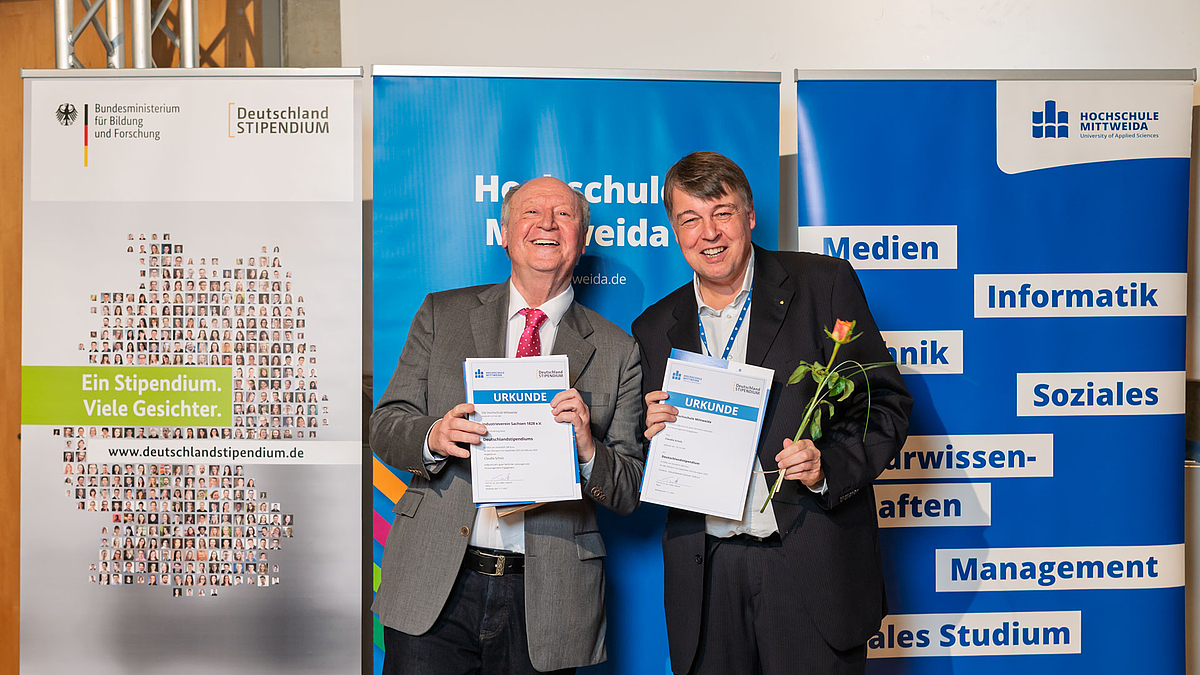 Nur die beiden übergenden Personen stehen lachend mit jeweils einer Urkunde vor den Rollups.