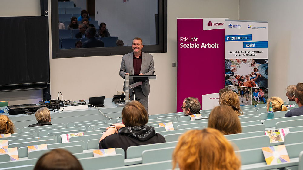 Männliche Person im Jackett lächelnd am Rednerpult im Hörsaal. Zwei Rollups im Hintergrund mit „Fakultät Soziale Arbeit“ und „Mittelsachsen Sozial“. Köpfe vom Publikum von hinten.