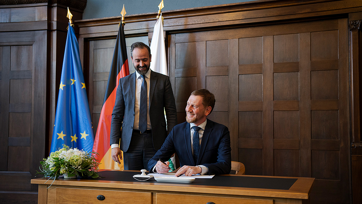Vor den Rektor:innen der sächsischen Hochschulen unterzeichneten Ministerpräsident Michael Kretschmer und Wissenschaftsminister Sebastian Gemkow die neue Zuschussvereinbarung. Ministerptäsident Michael Kretschmer sitzt einem Holzschreibtisch in der Sächsischen Staatskanzlei. Er grinst brei und hält einen Kugelschreiber in der rechten Hand.