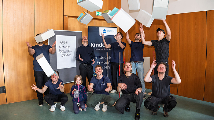 Eine Gruppe von zehn Personen im Hörsaal, die Schubladen aus Karton in die Luft werfen und wieder auffangen. Im Hintergrund sieht man ein dunkelblaues Kinderuni-Banner und eine Stellwand mit der Aufschrift „Neue Kinder kennenlernen“.
