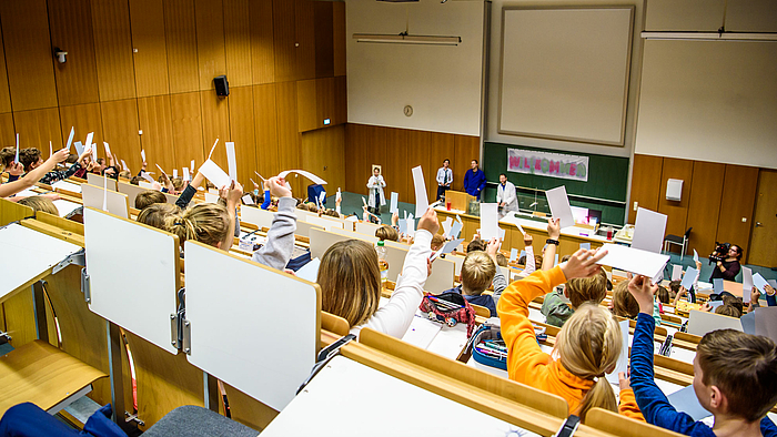 Das Foto zeigt von hinten einen großen Hörsaal mit aufsteigenden Sitzreihen. Der Hörsaal ist vollbesetzt mit Kindern, sie halten große Zettel nach oben. Das Foto blickt über die Köpfe und erhobenen Hände der Kinder nach vorne zum Pult, wo fünf weitere Personen zu sehen sind, teilweise in weißen Kitteln. 