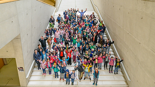 Grund zum Jubeln: Grundschule an der Hochschule. Die Schüler:innen aus Altmittweida waren zum Projekttag „Nachhaltigkeit“ an der Hochschule Mittweida. Blick von oben auf eine große Treppe. Viele Kinder und einige Erwachsene schauen mit hochgerissenen Armen jubelnd in die Kamera.
