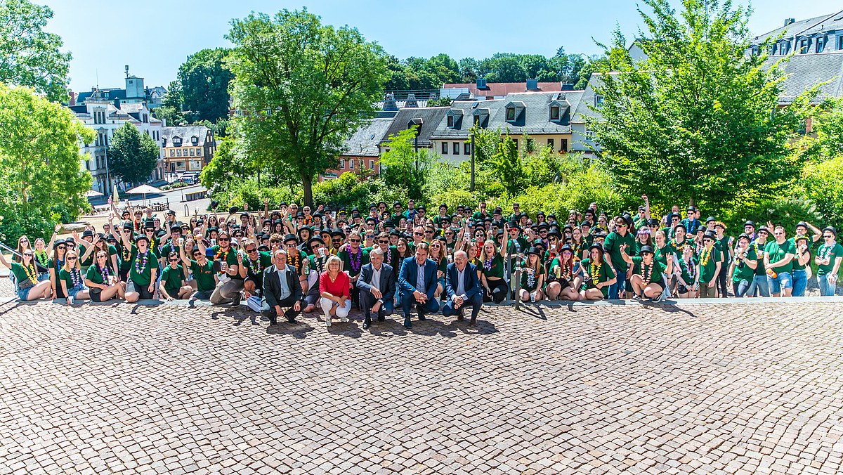 Das Gruppenoto zeigt zahlreiche übweiegen junge Menschen, viele mit Zylindern und einheitlichen T-Shirts. In der erstenten Reihe knien fünf ältere Personen. Alle schauen nach oben.