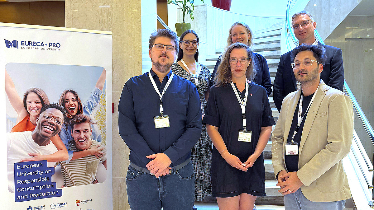 Die Hochschule Mittweida beteiligte sich durch vier Forschende an der Konferenz in Chania: (vorne v. l. n. r.) Felix Fischer, Anna Naumann, Dr. Markus Lohse sowie Dr. Jörg Schille (h. r.) Sechs Menschen stehen auf zwei Stufen einer Treppe und posieren förmlich für ein Gruppenfoto. Links im Bild steht ein Rollup mit einem Bild lachender, fröhlicher Menschen.