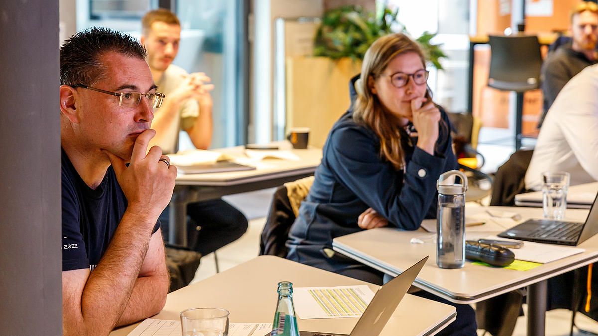 Eine männliche und eine weibliche Person sitzen jeweils an einem Tisch und schauen aufmerksam von ihren Laptops auf. Beide haben die rechte Hand konzentriert an ihren Mund gelegtIm Hintergrund sind unscharf weitere Personen zu sehen.