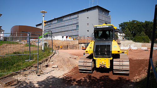 Anfang August 2025 begannen die Arbeiten für die Rohbau der neuen Hochschulbibliothek in Mittweida. Eine gelbe Planierraupe steht auf rotem Kiesboden. Im Vordergrund sieht man Bauzäune, im Hintergrund ein großes, modernes, graues Haus.