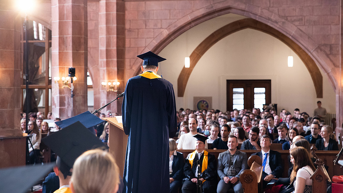 Professor Volker Tolkmitt spricht, den Rücken der Kamera zugewandt, zu den zahlreich auf den Kirchenbänken sitzenden Angehörigen und Studierenden.