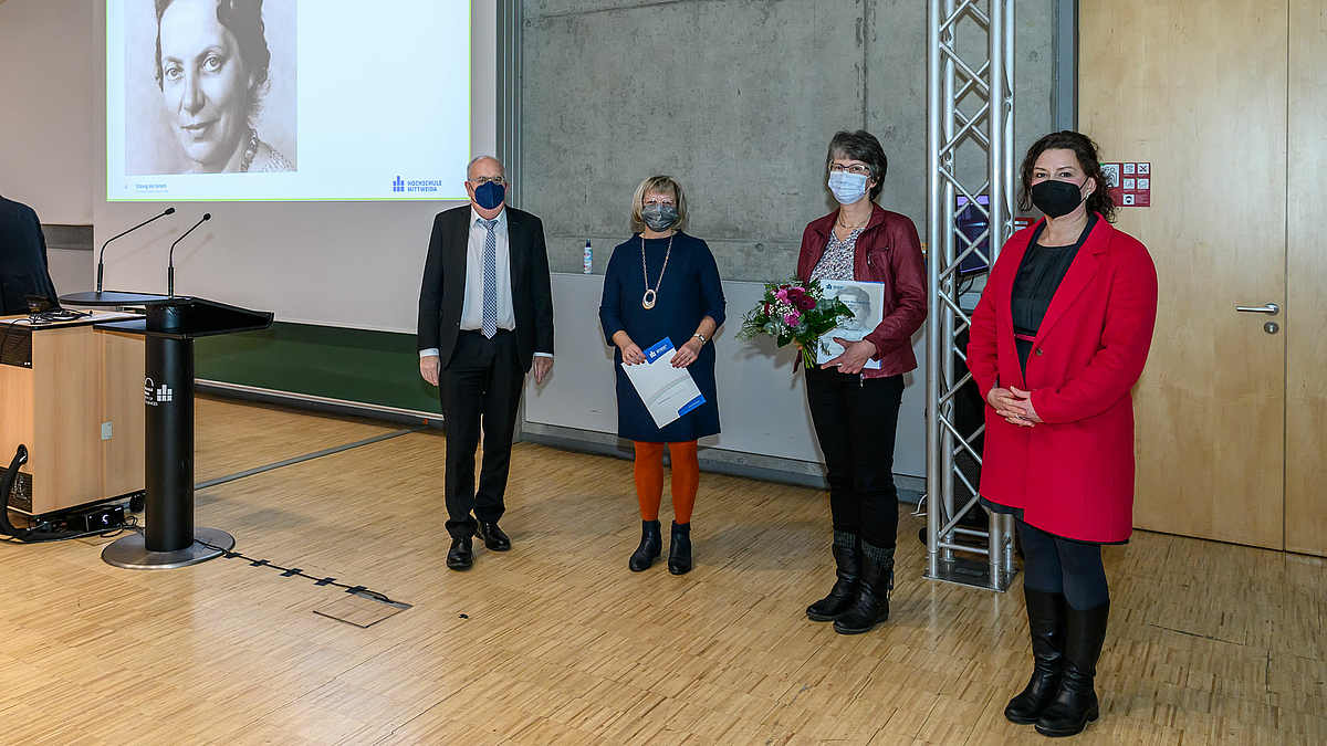 Auf dem Foto ist links auf dem Beamberbild das Porträt von Ingrid von Reyher zu sehen. in der erchen Bildhälfte sind vier stehende Personen mit Coronamaske zu sehen.