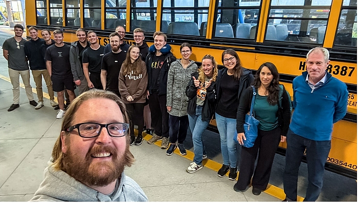 Mittweida goes Rochester. Professor Martin Anselm vom RIT begrüßt die frisch gelandete Gruppe der HSMW. Selfie zeigt eine Gruppe von überwiegend jungen Personen vor einem typischen gelben us-amerikanischen Schulbus. Im Bildvordergrund ist der Kopf des Fotograf zu sehen.